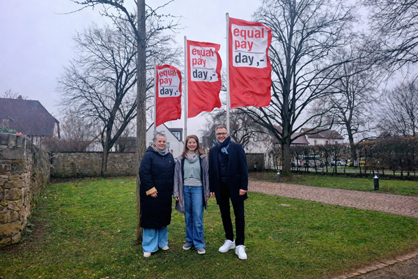 Die Fahnen zum Equal Pay Day sind vor dem Rathaus gehisst. V.l.: Katrin Roth und Johanna Fritz vom Frauenbüro und Bürgermeister Steffen Ball (Foto mit KI-Bearbeitung).